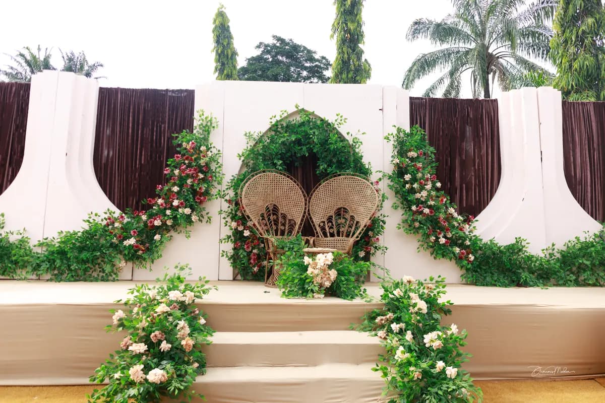 A traditional wedding stage designed as one set: two peacock chairs under a green arch, with roses and burgundy drapes.