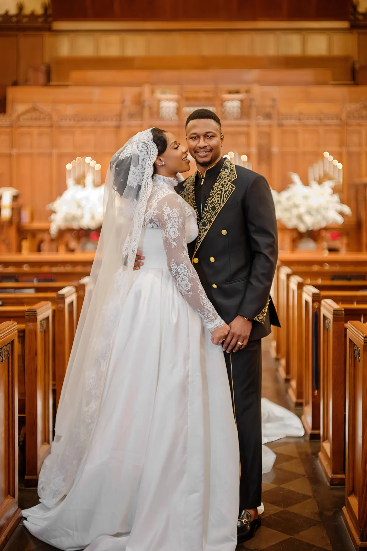 A bride and groom in the church aisle: her long-sleeved lace gown and veil, his black double-breasted suit with gold embroidery at the lapels.
